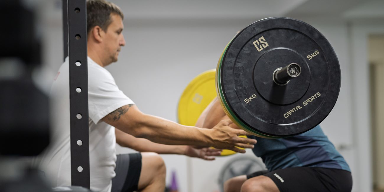 Guggolás elől tartott rúddal edzői korrekcióval a PiroskaGymben Bükön, technikajavítás közben.